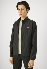 Donna che indossa una giacca zip-up nera di New Balance sopra una maglietta chiara, in piedi davanti a uno sfondo bianco semplice.