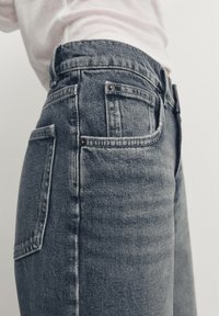 Gros plan d'une personne portant un jean en denim bleu délavé taille haute et une chemise blanche rentrée sur un fond uni.