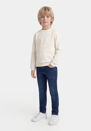 Lyshåret dreng står med den ene hånd i lommen og bærer en cremefarvet sweatshirt med præget logo, blå jeans og hvide sneakers på hvid baggrund.