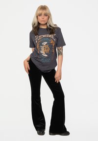 Gray graphic tee featuring Fleetwood Mac design, paired with black velvet flared pants. The outfit includes black boots and notable tattoos.