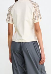 Kortärmad, krämfärgad topp med texturerade meshärmar och avslappnad passform. Matchad med lösa, grå byxor för en avslappnad stil.