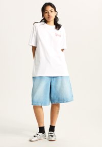 Hvid bomulds t-shirt med pink logo, parret med lyseblå denimshorts. Modellen har sorte sokker og sølvfarvede sneakers med velcro stropper på.