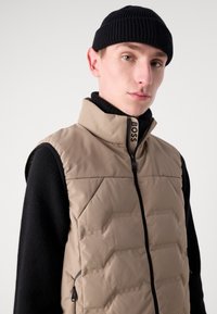 Beige quiltet vest med høy krage og glidelås, parret med en svart strikket genser. Merking på glidelåsknappen.