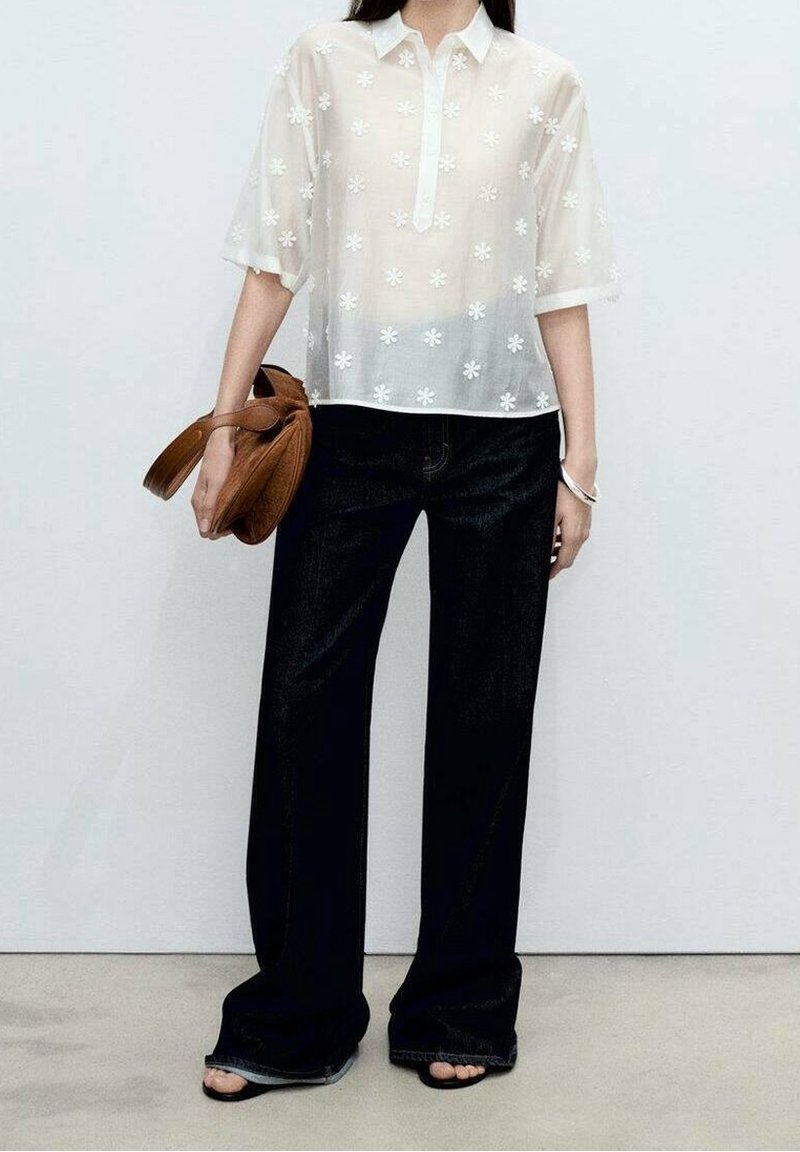 Femme portant un chemisier blanc transparent à motifs floraux et manches courtes, un pantalon noir à jambes larges, des sandales noires ouvertes aux orteils et tenant un sac à main marron.