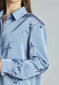 Camicia di raso azzurro chiaro con colletto, caratterizzata da ricami floreali in paillettes blu sulle maniche e dettagli arricciati ai polsini.