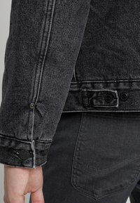 Black denim jacket with gray accents, featuring button closures on the cuffs and back, shown alongside dark pants with a pocket design.