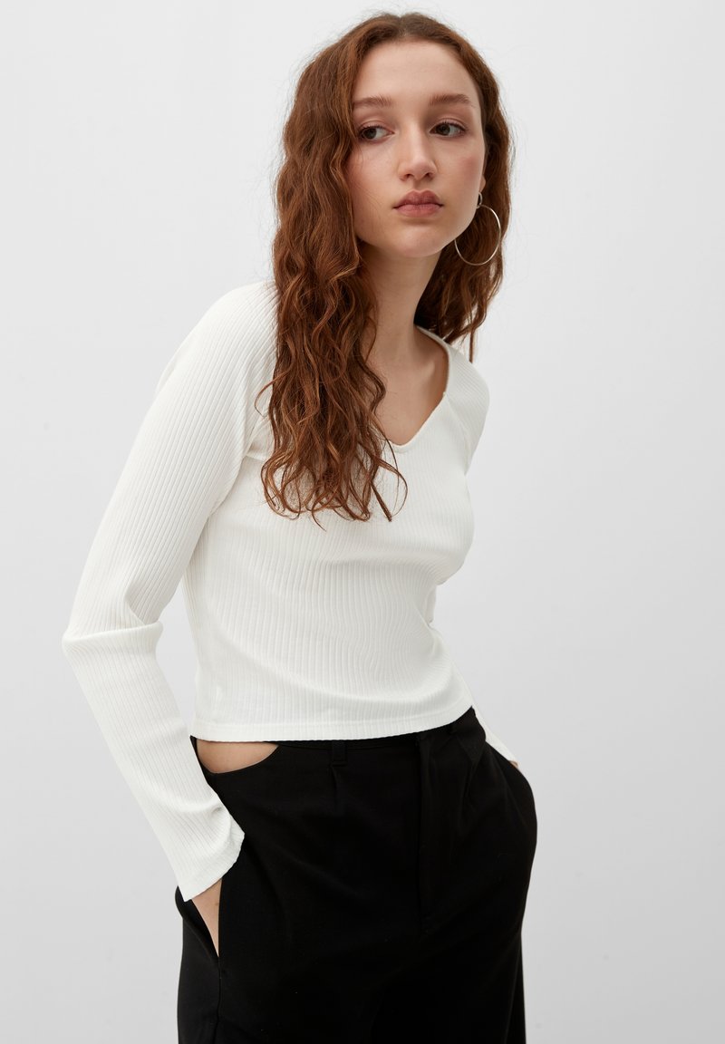 Witte ribbelde lange mouw top met een V-hals, gecombineerd met zwarte broek met zijzakken. De stof lijkt zacht en textuur te hebben.
