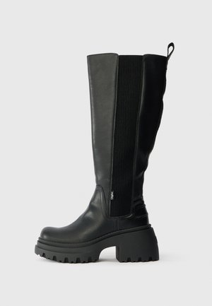 Black knee-high boots made of leather with a textured panel, a thick platform sole, and a pull tab at the back for easy wear.