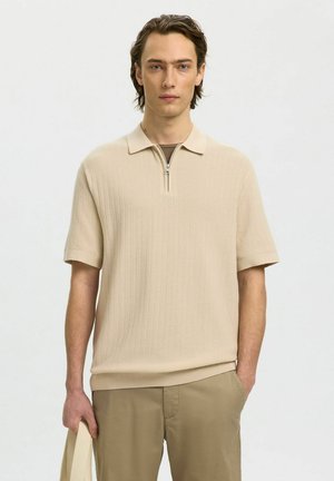 Giovane uomo con capelli castano chiaro che indossa una polo a maniche corte in maglia beige e pantaloni kaki, con in mano una borsa a tracolla beige.