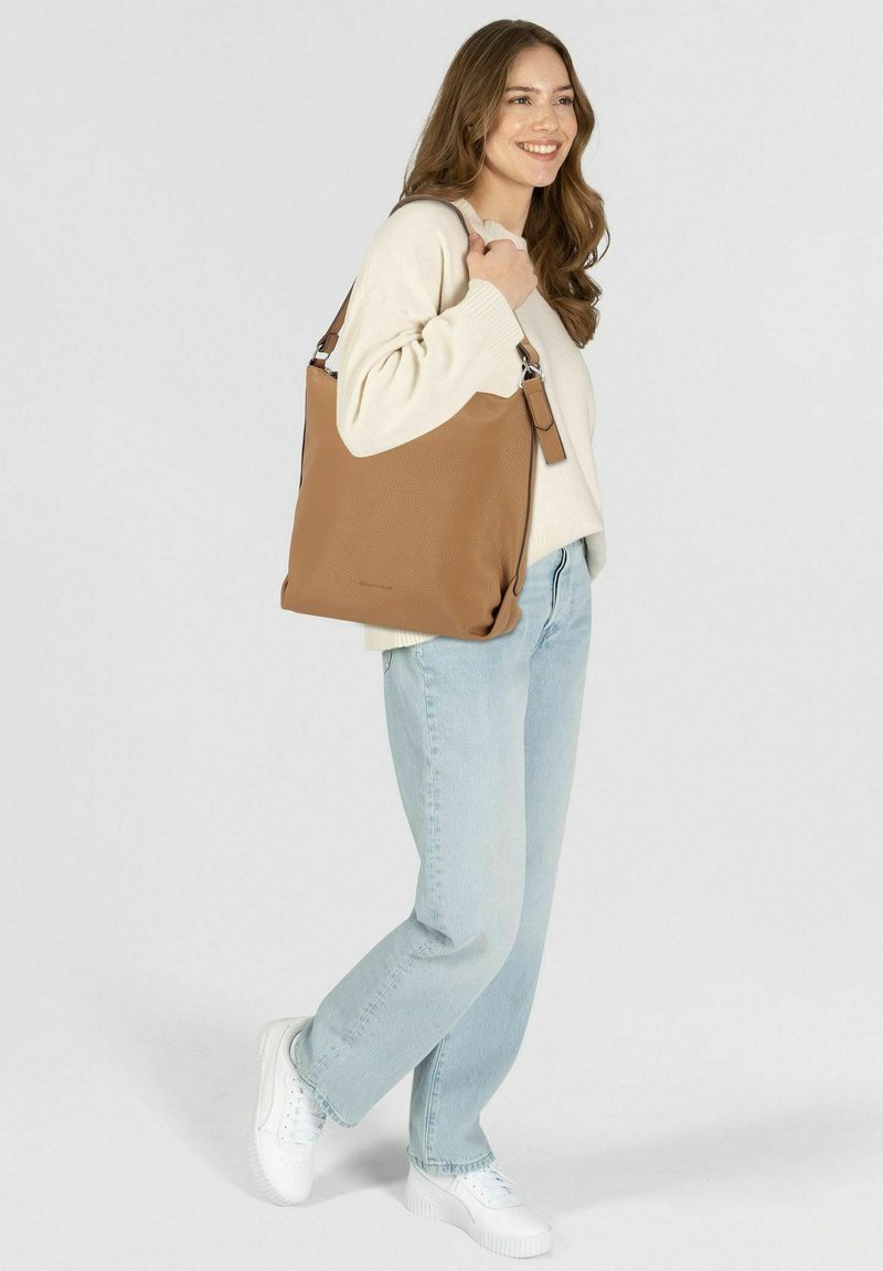 Braune Leder-Tote-Bag mit strukturiertem Finish, ausgestattet mit einem Schultergurt und silbernen Hardware-Elementen. Das Modell posiert lässig in hellen Jeans.