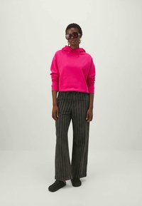 Woman wearing bright pink hoodie, black pinstriped wide-leg pants, black slip-on shoes, oversized sunglasses, and gold hoop earrings.