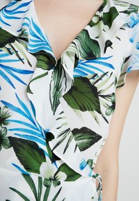 Blusa bianca con stampa di foglie tropicali in verde e blu, con scollatura arricciata e dettaglio di legatura laterale. Tessuto morbido e leggero.