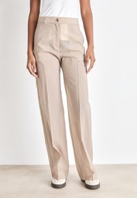Pantaloni beige a gamba larga con una leggera texture stropicciata, vita alta e un unico bottone frontale. Abbinati a sneakers di colore chiaro.