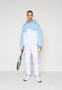 Chándal deportivo azul y blanco con capucha, que presenta un cierre frontal de cremallera, puños elásticos, y una raqueta y pelota de tenis. Tejido texturizado.