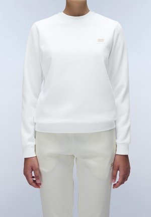 Persoon die een witte longsleeve crewneck trui en lichte broek draagt, staand tegen een eenvoudige, lichte achtergrond.