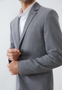 Grau suit Jackett aus strukturiertem Stoff, mit einem geknöpften Revers, einem einzelnen Knopfverschluss und einer Brusttasche. Darunter ein weißes Hemd mit Kragen.