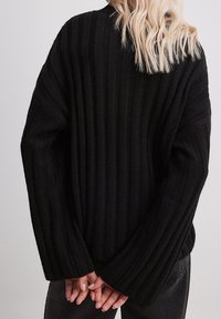 Pull noir à texture côtelée avec manches longues et larges poignets, présentant une coupe ample. La vue de dos montre des lignes épurées et un design simple.