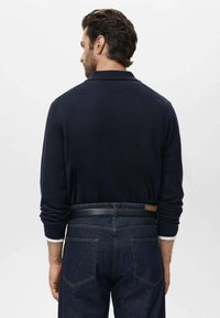 Suéter de polo de punto azul marino con diseño de cuello, combinado con jeans de mezclilla oscura y un cinturón negro, cuenta con puños ajustados y una textura suave.