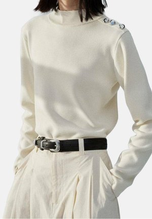 Woman wearing cream long-sleeve sweater with shoulder buttons, tucked into high-waisted light pants with a black belt, hands in pockets.