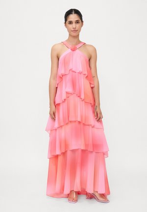 Pink tiered maxi dress with flowing chiffon fabric. Features a rose accent at the neckline and layered ruffles. Transparent heels are worn.