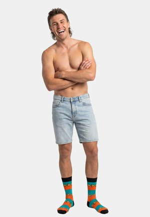Lächelnder, oberkörperfreier Mann mit verschränkten Armen, trägt hellblaue Jeansshorts und bunte gemusterte Socken, steht vor einem einfarbigen Hintergrund.