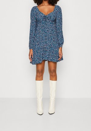 Kobieta w niebieskiej, kwiecistej, długorękawowej mini sukience, zestawionej z białymi kozakami za kolano, stojąca na tle jasnego, jednolitego tła.