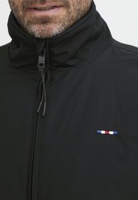 Črna jakna z zadrgo in visokim ovratnikom, ki ima majhen vezen rdeč, bel in moder vodoraven trak na prsih. Gladek tekstura tkanine.