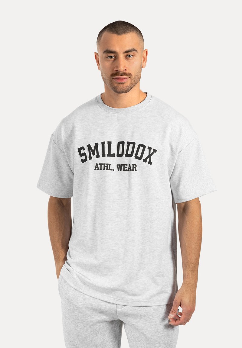 Camiseta gris de corte oversize hecha de una mezcla de algodón, con un texto impreso en negro "SMILODOX ATHL. WEAR" en el frente. Manga corta y ajuste relajado.