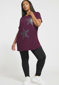 Nainen, jolla on yllään violetti yli suuri t-paita, jossa on kaksi kuvioitua tähteä, mustat leggingsit ja valkoiset lenkkarit, poseeraa yksinkertaista valkoista taustaa vasten.