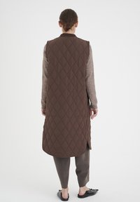 Vadderad brun väst med diamantmönster, ärmlös design, sidofickor, kombinerad med grå-bruna byxor och svarta platta skor.