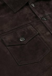 Bruin suède stof shirt met een scherpe kraag, voorzien van een borstzak en zilveren drukknoopsluiting. Zachte textuur met subtiele glans.