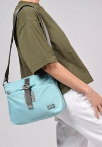 Sac à épaule synthétique bleu clair avec des accents gris, comprenant une poche avant et une bandoulière ajustable, porté de manière décontractée sur une chemise verte.