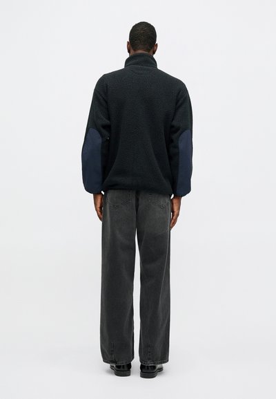 Μαύρος φλις πόλο με ψηλό γιακά; οι μανίκες διαθέτουν ναυτικά υφασμάτινα αξεσουάρ. Συνδυάζεται με φαρδιά γκρι τζιν και μαύρα παπούτσια.