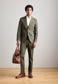 Homme portant un costume vert olive et une chemise blanche, tenant un sac en cuir marron, se tenant sur un sol en bois contre un mur gris clair.