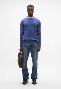 Lila Pullover mit Rundhalsausschnitt, kombiniert mit blauen Jeans. Das Model hält eine braune Lederhandtasche und trägt dunkle, klobige Schuhe.