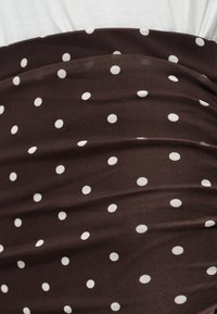 Brązowa tkanina z równomiernie rozmieszczonymi białymi kropkami, lekko pognieciona, na jednolitym jasnym tle.