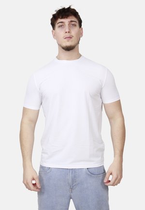 Giovane uomo con capelli corti e ricci che indossa una t-shirt bianca semplice a maniche corte e jeans azzurri, in piedi contro uno sfondo chiaro.