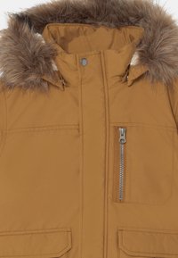 La doudoune marron présente une capuche doublée en fausse fourrure, une poche zippée à l'avant et deux grandes poches frontales avec fermetures à pression. La texture est en tissu lisse.