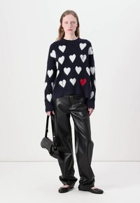 Maglione blu navy con motivi di cuori bianchi e rossi, abbinato a pantaloni in pelle neri e scarpe basse nere, portava una piccola borsa nera.