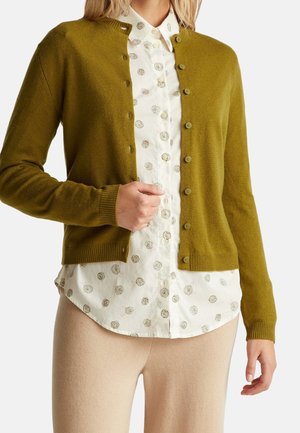 Femme portant un cardigan boutonné vert olive sur une chemise blanche à fleurs boutonnée et un pantalon beige, debout devant un fond blanc.