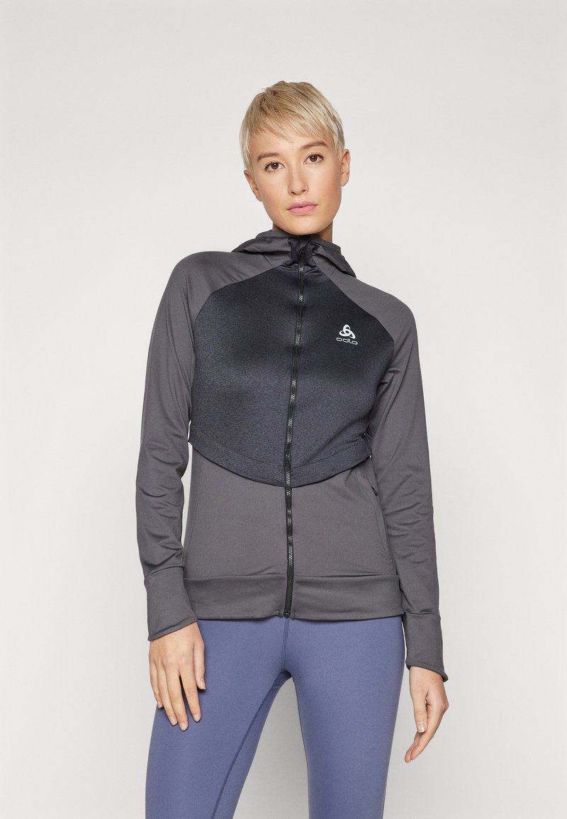 Sportjacke in Dunkelgrau mit Reißverschluss vorne, ausgestattet mit einem strukturierten Brustpanels, Kapuze und figurschmeichelndem Design. Kombiniert mit blauen Leggings.