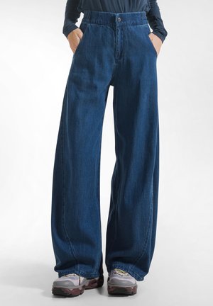 Persoon draagt losse donkerblauwe denim jeans met handen in de zakken en grijze sneakers met bordeauxkleurige zolen.
