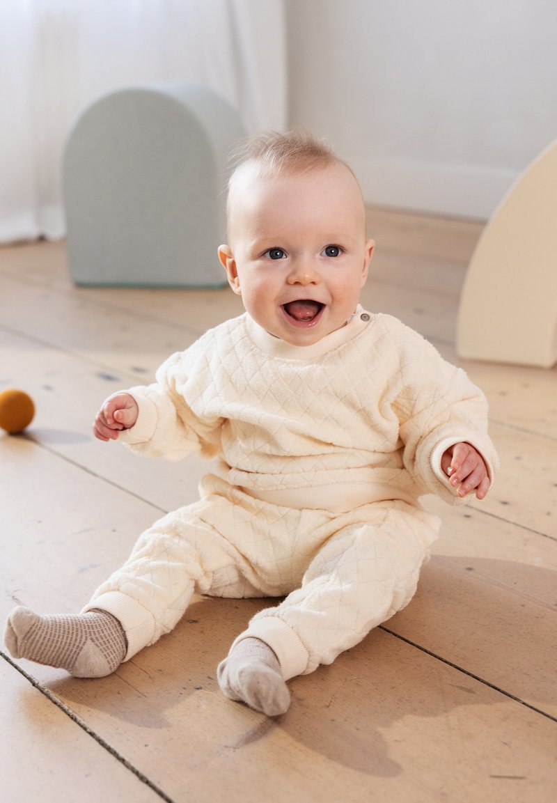 Beige, gesteppter Babyanzug mit elastischen Bündchen, der auf einem Holzboden sitzt. Auffällige Texturen und ein Knopfdetail an der Schulter.