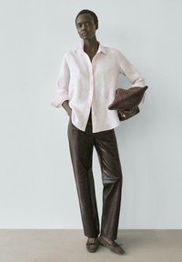 Žena v světle růžové košili na knoflíky, tmavě hnědých kožených kalhotách, drží tkané hnědé psaníčko, stojí před jednoduchou světlou zdí.