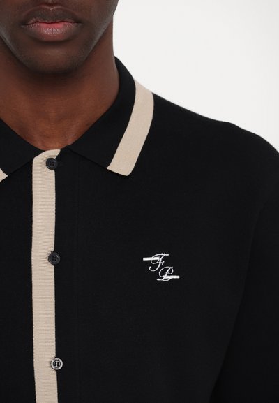 Schwarzes Poloshirt mit einem beigen Streifen entlang des Kragens und der Knöpfe. Verfügt über ein gesticktes Logo auf der linken Seite. Der Stoff hat eine glatte Textur.