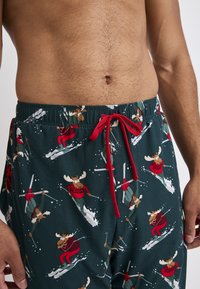 Groene pyjamabroek met een speels patroon van skiënde elanden, voorzien van een rode trekkoord en een elastische tailleband. Het materiaal lijkt zacht en lichtgewicht te zijn.