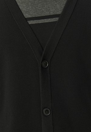 Zwarte cardigan met een v-hals, knoopsluiting en zachte brei-textuur. Gelaagd over een grijze shirt, met een gestreept patroon eronder.