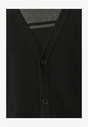 Zwarte cardigan met een v-hals, knoopsluiting en zachte brei-textuur. Gelaagd over een grijze shirt, met een gestreept patroon eronder.