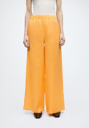 Žena stojaca na jednoduchom pozadí, oblečená v oranžových nohaviciach s vysokým pásom a širokými nohavicami, s elastickým pásom a čiernymi topánkami so štvorcovou špičkou.