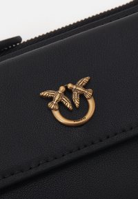 Bolsa de couro texturizado preto com uma emblema em metal dourado de duas aves circundando um anel. Detalhes de costura sutis ao longo das bordas.